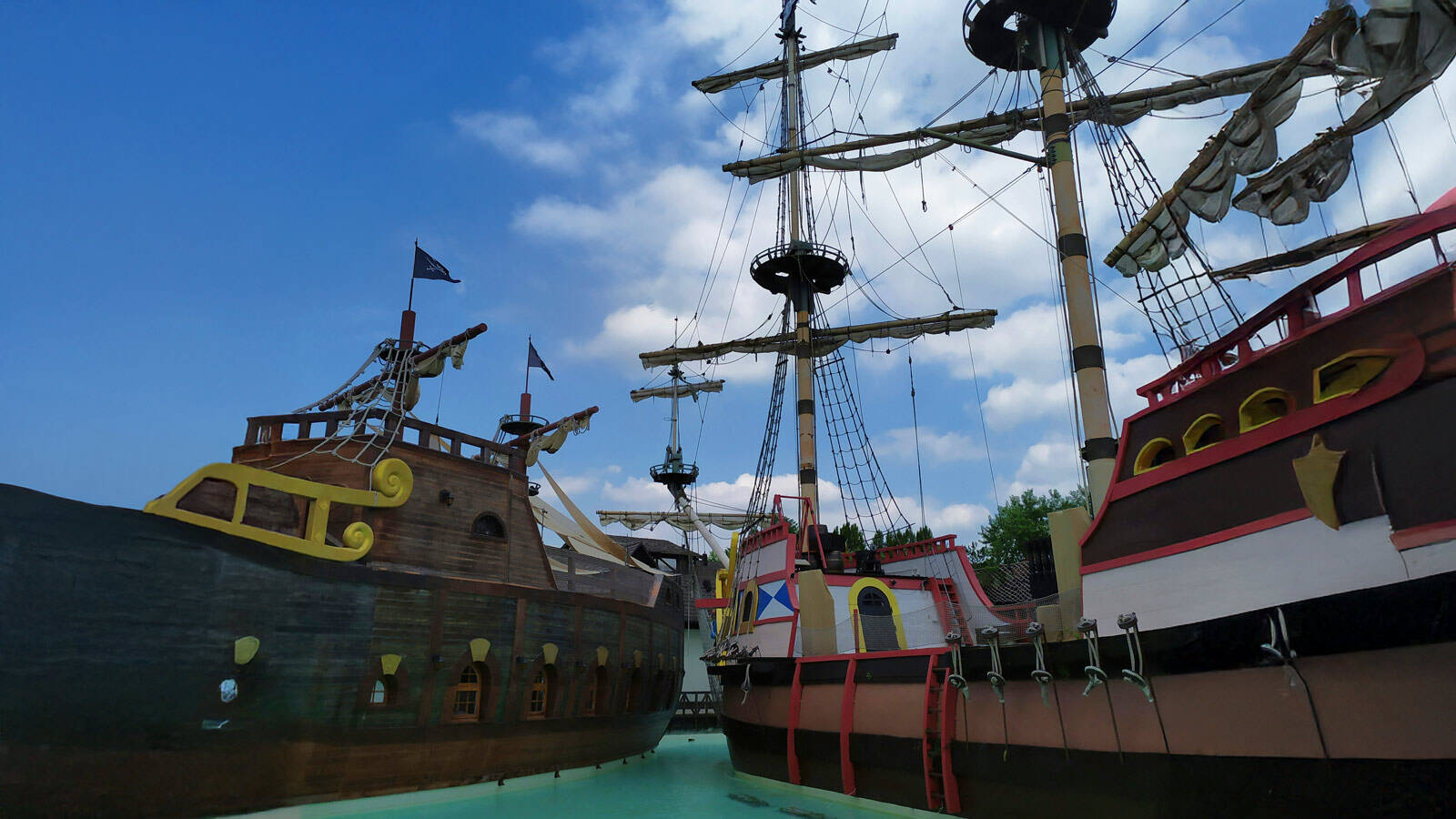Des reconstitutions de galions ou navires de pirates dans le parc d'attractions Gardaland, avec leurs mâts et voiles, sur un plan d'eau, sous un ciel bleu parsemé de nuages blancs.